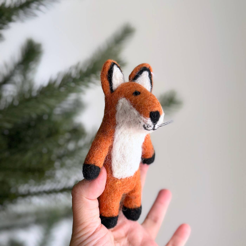 A hand holds up a Felt Finger Puppet: Fox in front of blurred green pine branches and a light background—ideal for storytelling.
