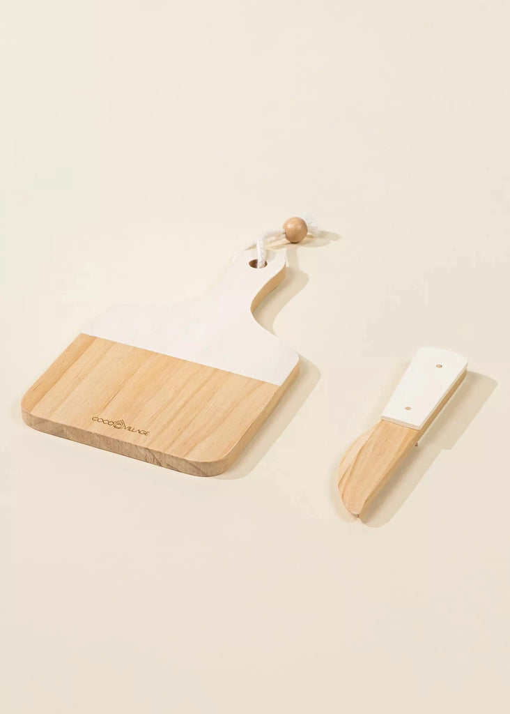 The Wooden Bakery Set, featuring a rectangular wooden cheese board with a white painted top half and a hanging string loop, is displayed alongside a small matching wooden and white cheese knife. This set is ideal for enjoying various types of bread and both items are placed on a light beige background.