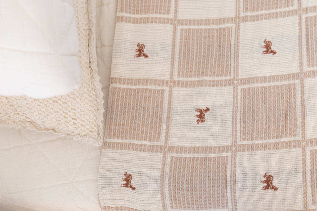 A close-up of the Organic Cotton Twin Size Blanket - Pony, featuring a beige and white checkered design with red pony motifs, draped over a bed with a cream organic cotton pillow and delicate crochet trim.
