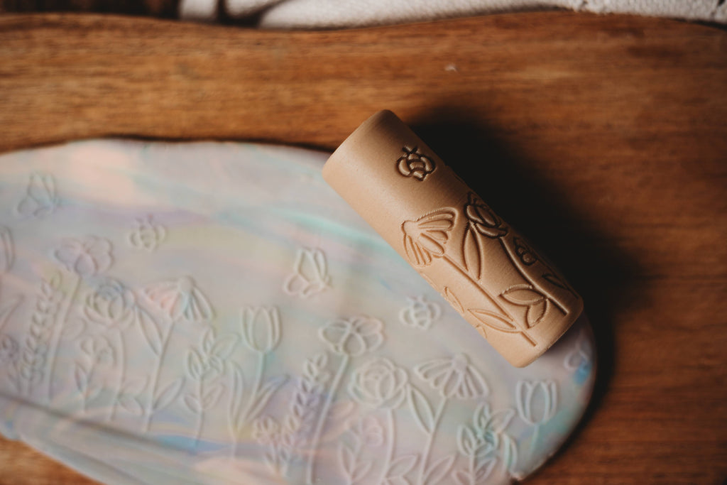 The Flower Roller - Eco Pattern Playdough Roller features elegant floral engravings and is shown resting on a pastel marbled clay slab embossed with a matching flower design, all set atop a wooden surface—making it perfect for sensory play and an ideal addition to your collection of eco-friendly playdough tools.