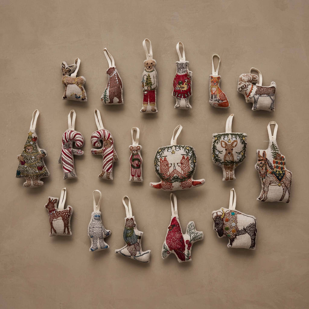 A Coral & Tusk Red Cardinal Ornament among a collection of seventeen assorted embroidered fabric ornaments on natural linen, featuring animals, candy canes, and holiday themes.