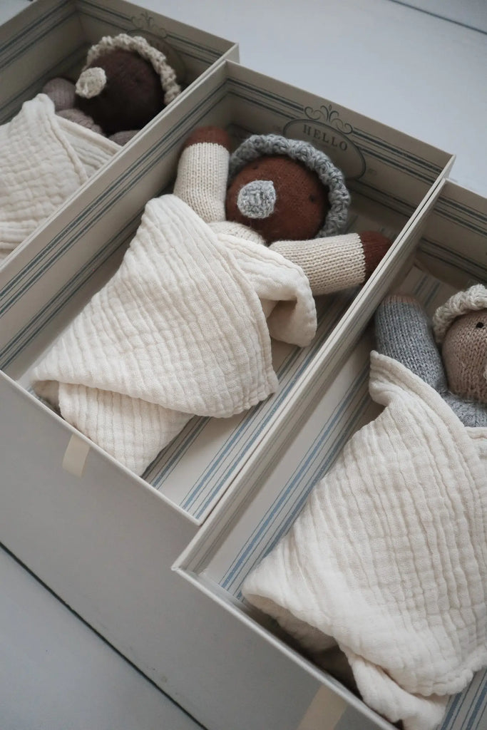 Three Hand-Knit Baby Doll - Cocoa shaped like bunnies, each wrapped in an organic cotton swaddle, are nestled in separate compartments of a wooden box with a "hello" greeting on one compartment's edge.