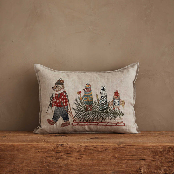 The Coral & Tusk Christmas Tree Sled Pillow is a rectangular beige holiday pillow featuring embroidered animals in winter clothes as a bear pulls a sled with a tree and three animals, set against a plain brown wall.