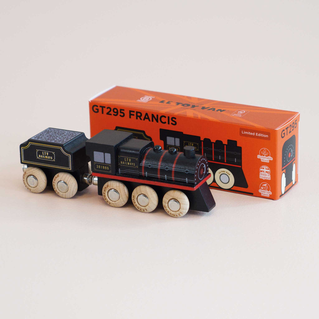 A black wooden toy train engine with gold and red details sits in front of its orange box labeled “No. 49 GT295 Francis | Made in England” and “Limited Edition.” Inspired by classic locomotives, this steam train toy features white wheels, while the box showcases an illustration of the train.