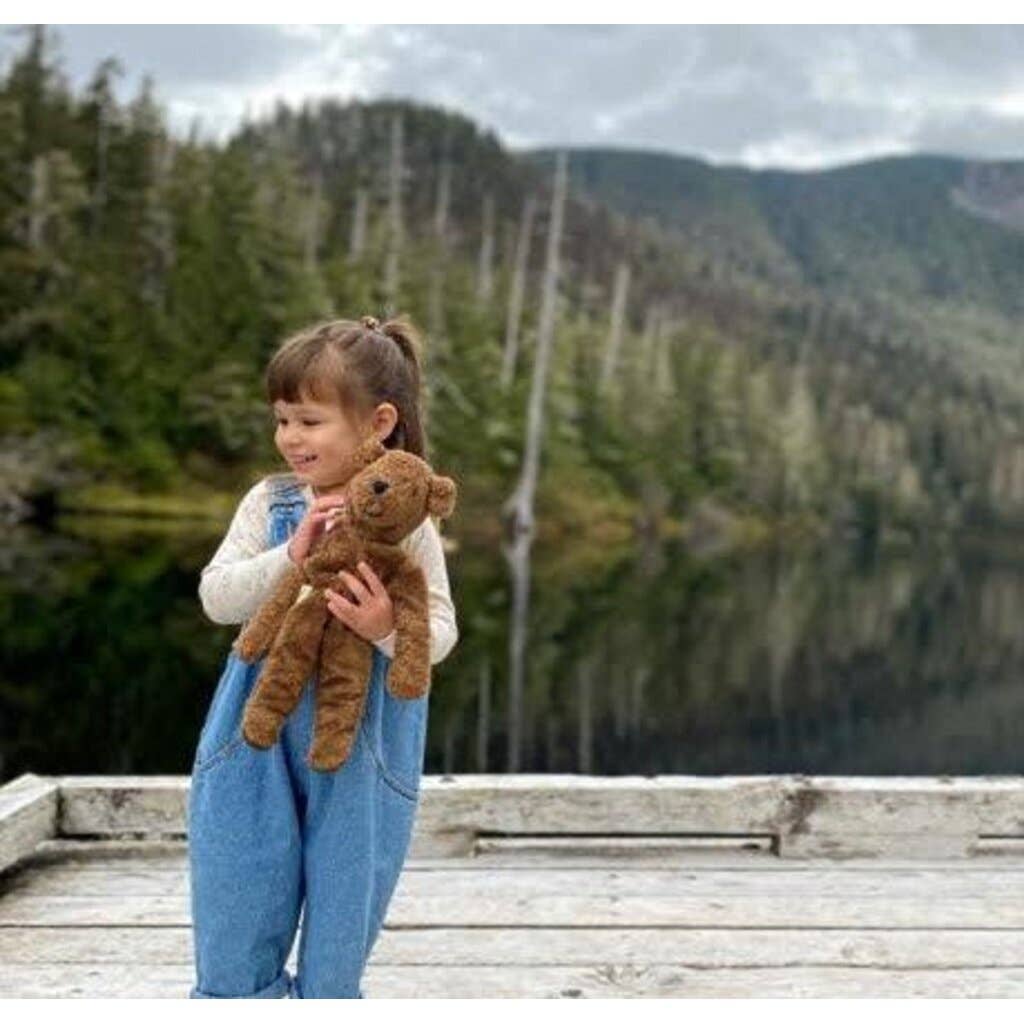 A young girl in blue overalls holds a Senger Naturwelt Stuffed Animal - Large Brown Bear while standing on a wooden dock by a calm lake, with evergreen trees and mountains in the background.