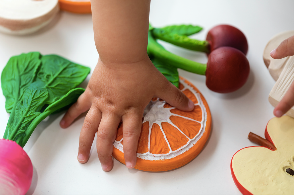 A child’s hand touches Clementino the Orange Teether, surrounded by other colorful wooden toy fruits and vegetables on a white surface—perfect for sensory play.