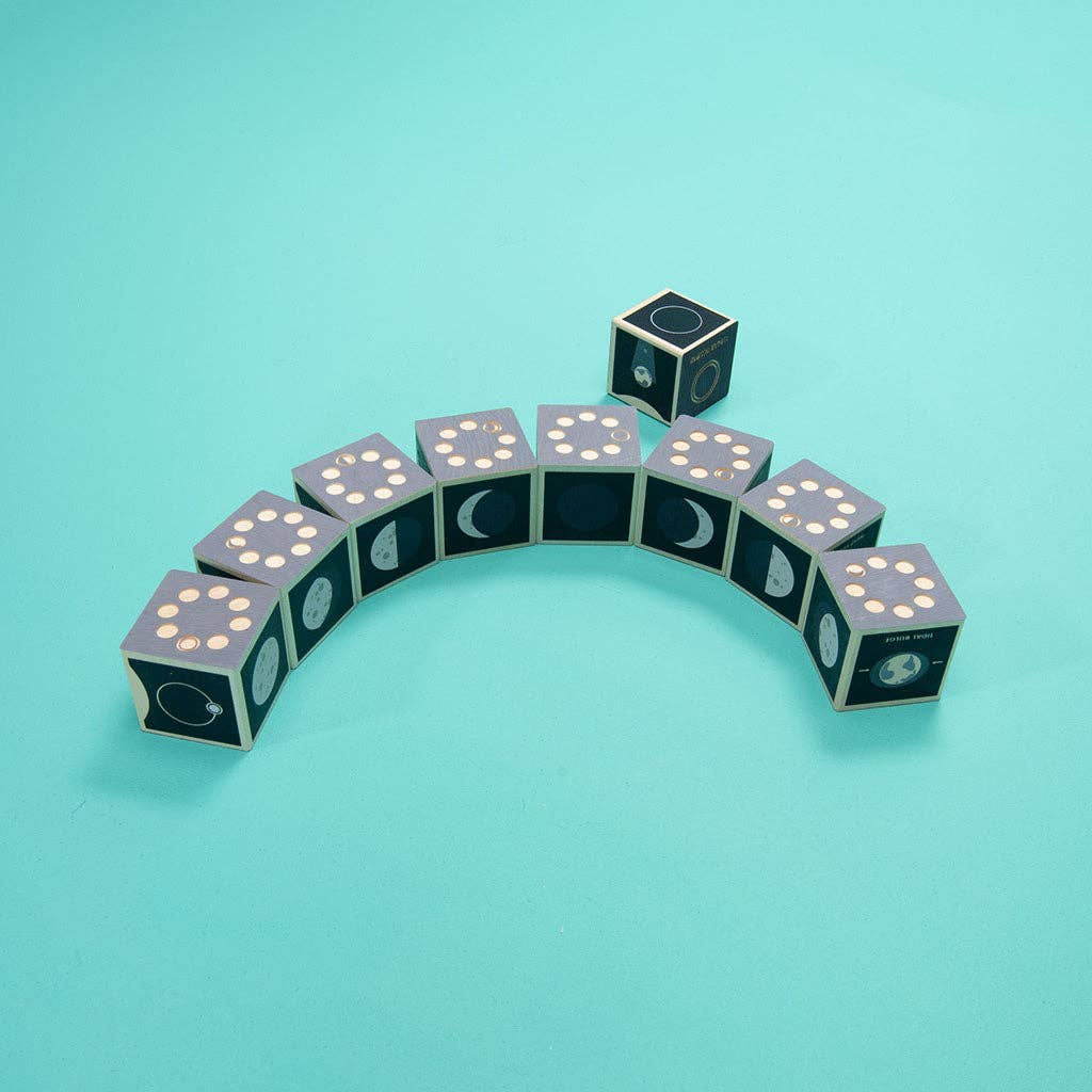 Nine Uncle Goose Moon Phase Blocks are arranged in an arc on a teal surface, each featuring a different phase of the moon and white dice-like dots. One block is set apart above the arc, highlighting the changing beauty of the moon’s phases.