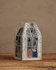 The Coral & Tusk Haunted House Tissue Box Cover features arched windows, spiderwebs, and embroidered Halloween characters—a skeleton, witch, and pumpkin—peeking out atop a wooden surface.