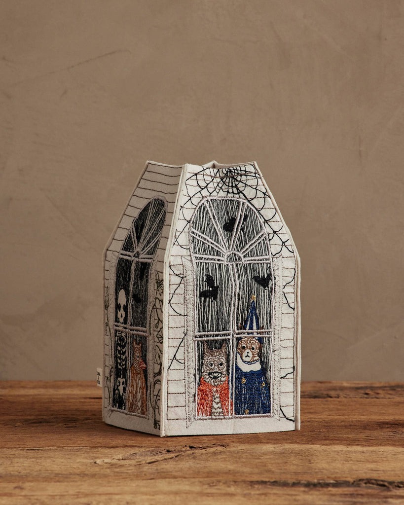 The Coral & Tusk Haunted House Tissue Box Cover features arched windows, spiderwebs, and embroidered Halloween characters—a skeleton, witch, and pumpkin—peeking out atop a wooden surface.