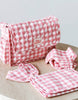 The Tiny Tummies Pink Gift Box - The Essentials by Tiny Harlow is showcased on a wooden surface, featuring a pink and white checkered quilted diaper bag ideal for doll essentials. Complementing the bag are matching accessories: a bib, a changing mat, and a cloth diaper. The "tiny harlow" brand label is prominently displayed on the bag.