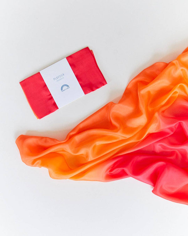 A folded Sarah's Silks Enchanted Playsilk - Fire, with a red and orange gradient and white label, lies beside a larger matching piece—ideal for open-ended play—on a white background.