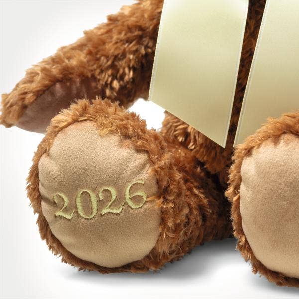 A close-up of the Steiff Cosy Year Bear 2026 13″ shows "2026" embroidered in yellow on its left paw. Finished with a cream ribbon around its neck, this bear makes a perfect keepsake or New Year’s gift.