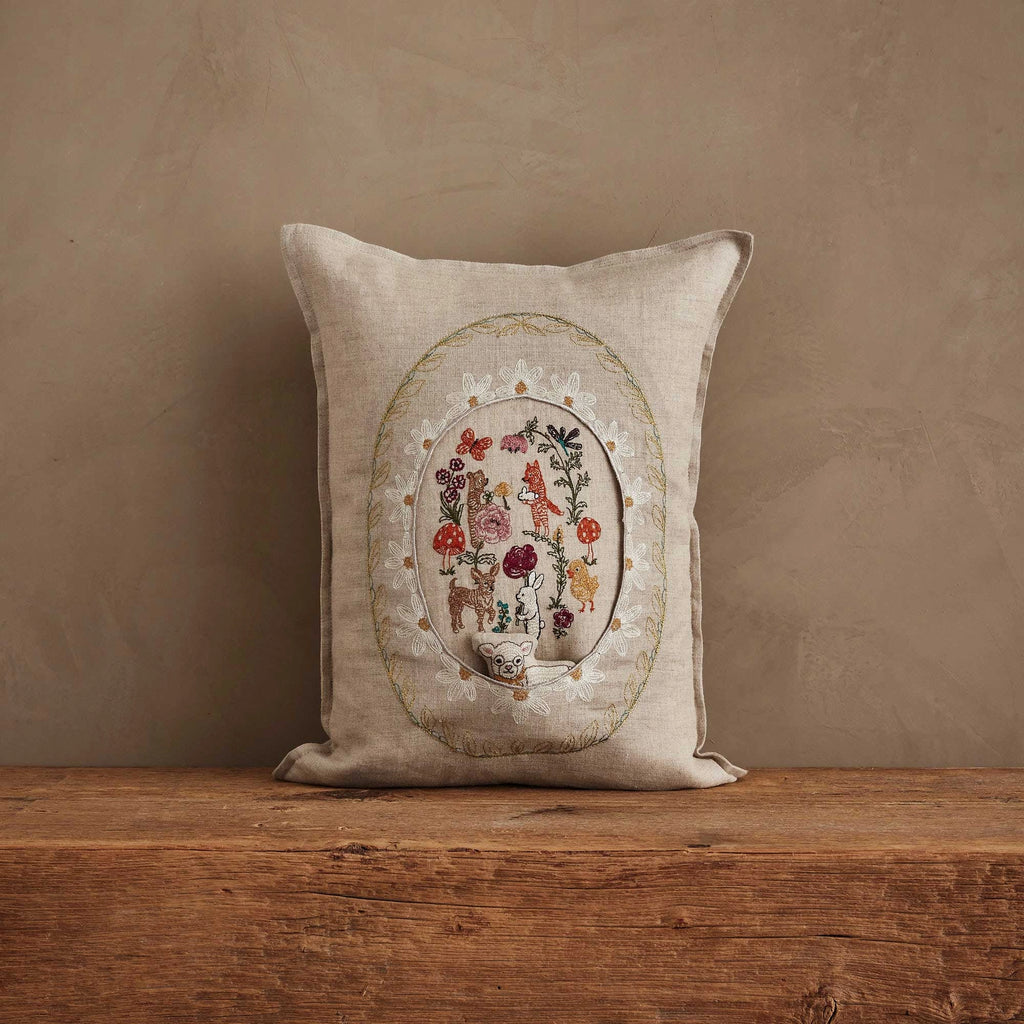 The Coral & Tusk Sugar Egg Pocket Pillow features embroidered florals and animals in an oval frame, creating a charming Easter accent on a rustic wooden surface against a textured beige wall.