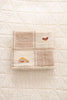 The Organic Cotton Baby Blanket - Farm, in beige and cream patchwork with embroidered chickens and a yellow house, is folded on a quilted off-white surface.
