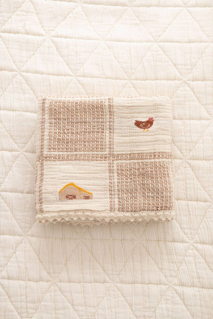 The Organic Cotton Baby Blanket - Farm, in beige and cream patchwork with embroidered chickens and a yellow house, is folded on a quilted off-white surface.