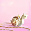 A close-up image of a Snail Necklace with intricate, realistic details hanging from a 24k gold plated chain. The background is a soft pastel pink, creating a charming and whimsical appearance that highlights its heirloom quality craftsmanship.