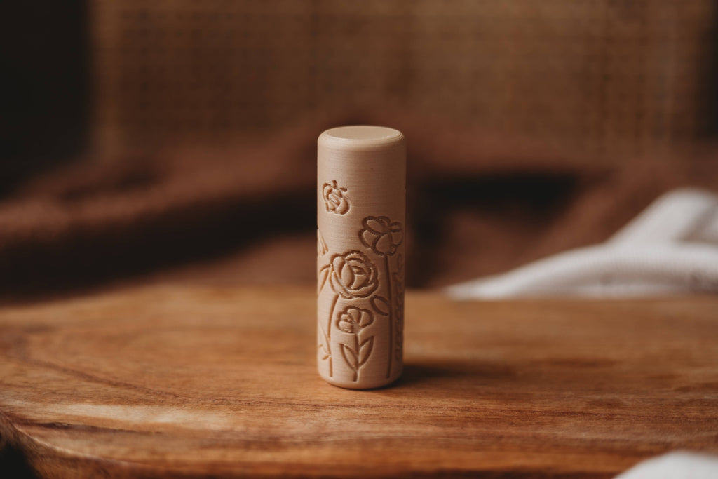The Flower Roller - Eco Pattern Playdough Roller, a cylindrical wooden stamp with intricate floral carvings, stands upright on a wooden surface against a soft brown backdrop—an ideal eco-friendly tool for sensory play and creative playdough activities.