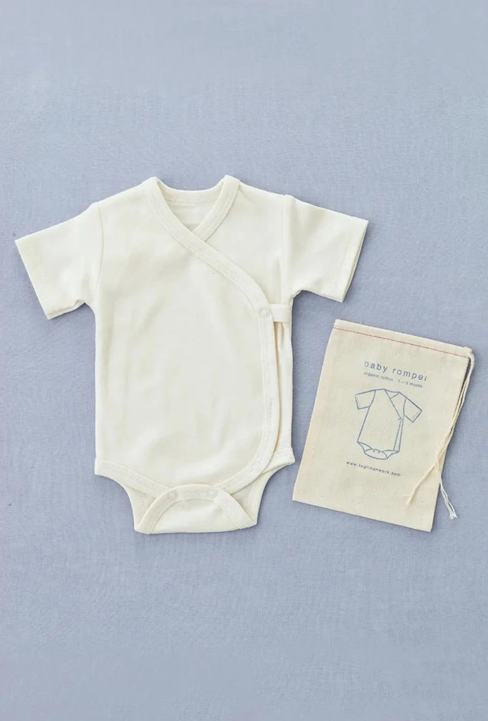 A cream-colored Baby Romper with a wrap-style front and snap buttons is placed on a light gray background. Beside it is a beige drawstring cloth bag with an illustration of a romper and the text "baby romper" printed on it. Machine wash for easy care.
