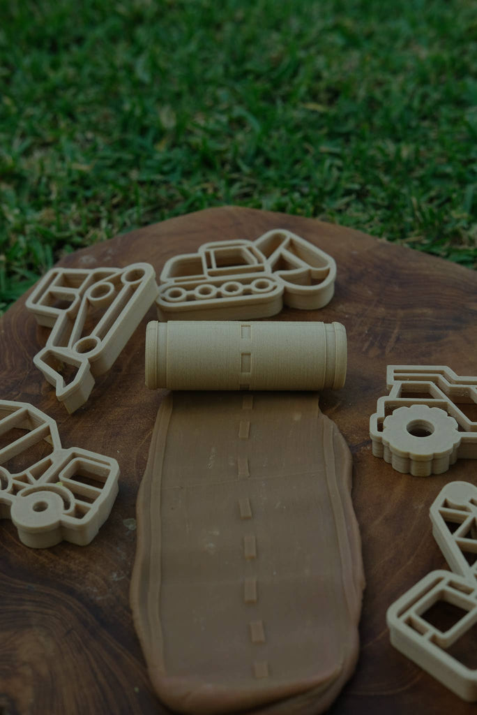 Toy construction vehicle-shaped cookie cutters and the Road Roller - Eco Pattern Playdough Roller are arranged on a board, with grass in the background. The eco-friendly Road Roller has imprinted a winding road pattern onto brown dough, making it perfect as a sensory play tool and for inspiring imaginative fun.