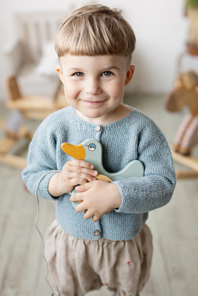 A smiling young boy holding a Mint Green Duck Pull Toy, wearing a blue sweater and beige pants, standing in a warmly decorated room with wooden toys in the background.