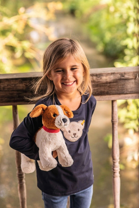 A joyful young girl stands on a wooden porch, holding a Steiff Snuffy Dog Plush Puppy, 11", wearing a dark blue top with a puppy design. blurred greenery in the background.