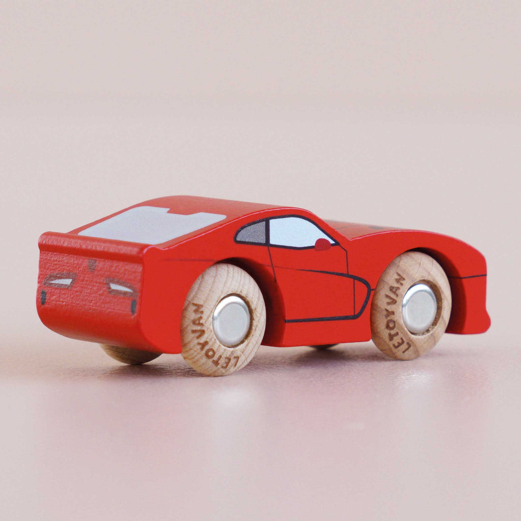 The No. 18 GT100 Jasper, handmade in England from sustainable wood, sits atop a pale pink surface. This small red wooden sports car features wooden wheels, simple cartoon-inspired details, and black-painted windows and doors—a playful nod to classic performance motoring.