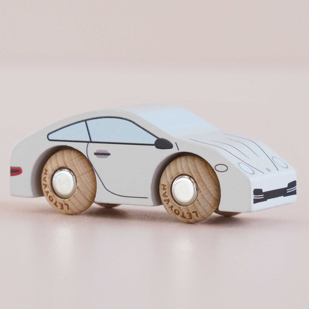 No. 16 GT90 Poppy is a wooden sports car painted white with black and blue accents, featuring simple wooden wheels. Its design is minimalist and stylized, reminiscent of a classic toy car, and it is photographed against a light pink background.
