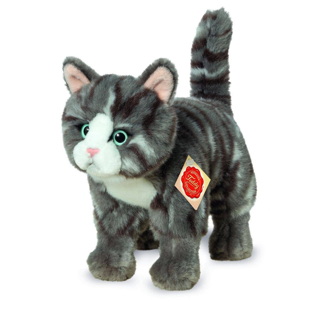 Introducing the Teddy Hermann Standing Tabby Cat Stuffed Animal, a high-quality plush toy cat with gray fur adorned with darker stripes, white accents on its face, chest, and paws, and striking green eyes. This cuddly cat is in a standing position with its tail upright and features a red tag with gold trim attached to its collar.