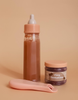 A Tiny Tummies Puree and Milk Bottle set for dolls, featuring a baby bottle filled with chocolate milk, a small jar labeled "Tiny Tummies chocolate puree," and a peach-colored baby spoon, are arranged against a beige background.