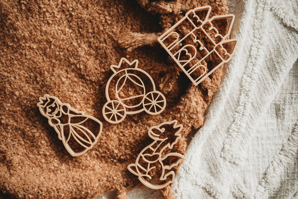The Princess Cutter Set - Eco Playdough Tool features four eco-friendly wooden cutters in the shapes of a princess, carriage, dragon, and castle, beautifully arranged on textured brown and white fabric—an ideal set for inspiring creativity during princess-themed play.