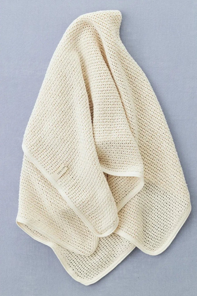 A soft, cream-colored, organic cotton Knit Baby Blanket is draped on a blue surface. The blanket features a delicate, mesh-like pattern and neatly finished edges.