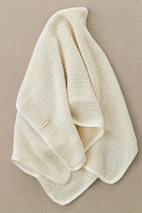 The Knit Baby Blanket, crafted from luxurious Lithuanian cotton in a cream-colored, textured knit, drapes elegantly against a beige background, highlighting its cozy and delicate fabric.