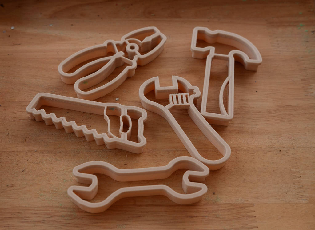 Five beige plastic cookie cutters shaped like hand tools—a saw, pliers, wrench, adjustable wrench, and hammer—are arranged on a wooden surface. The Tools Cutter Set - Eco Playdough Tool adds creative and sustainable fun to crafting sessions with its charming tool-shaped cutter designs.