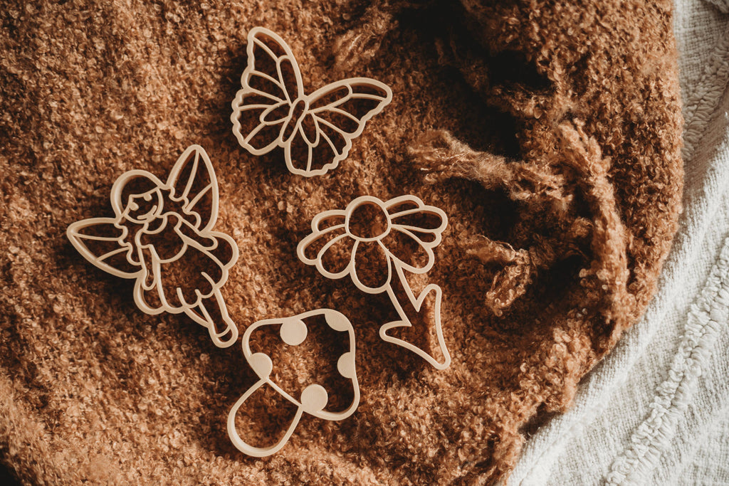 The Fairy Cutter Set - Eco Playdough Tool features four cookie cutters shaped like a fairy, a butterfly, a flower, and a mushroom displayed on textured brown fabric—creating the perfect fairytale cutter set for imaginative sensory play.