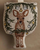 The Coral & Tusk Togetherness Deer Ornament features a brown and white fawn beneath a green pine wreath adorned with yellow and silver baubles, all on a beige fabric background.