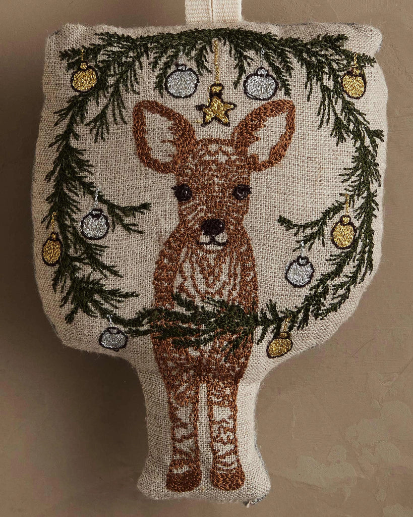 The Coral & Tusk Togetherness Deer Ornament features a brown and white fawn beneath a green pine wreath adorned with yellow and silver baubles, all on a beige fabric background.