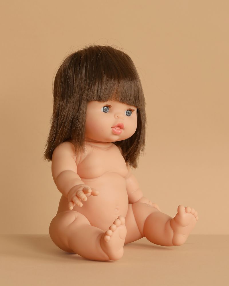 The Minikane Baby Doll (13") - Chloe, made in Spain, is an anatomically correct nude doll with shoulder-length brown hair, bangs, and blue eyes. The doll sits with arms and legs slightly forward on a beige surface. Clothing sold separately.