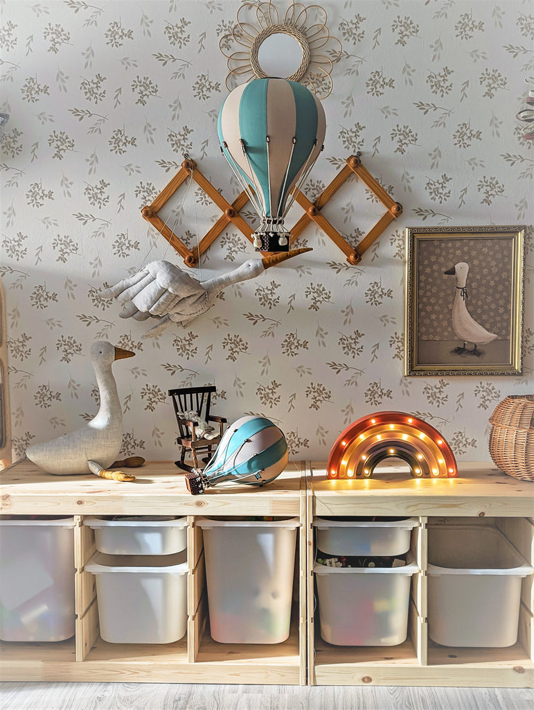 A children's room with wooden storage cubes, colorful bins, the Hot Air Balloon - Pastel Green decoration, a goose statue, a rainbow-shaped light, and framed artwork set against floral wallpaper.