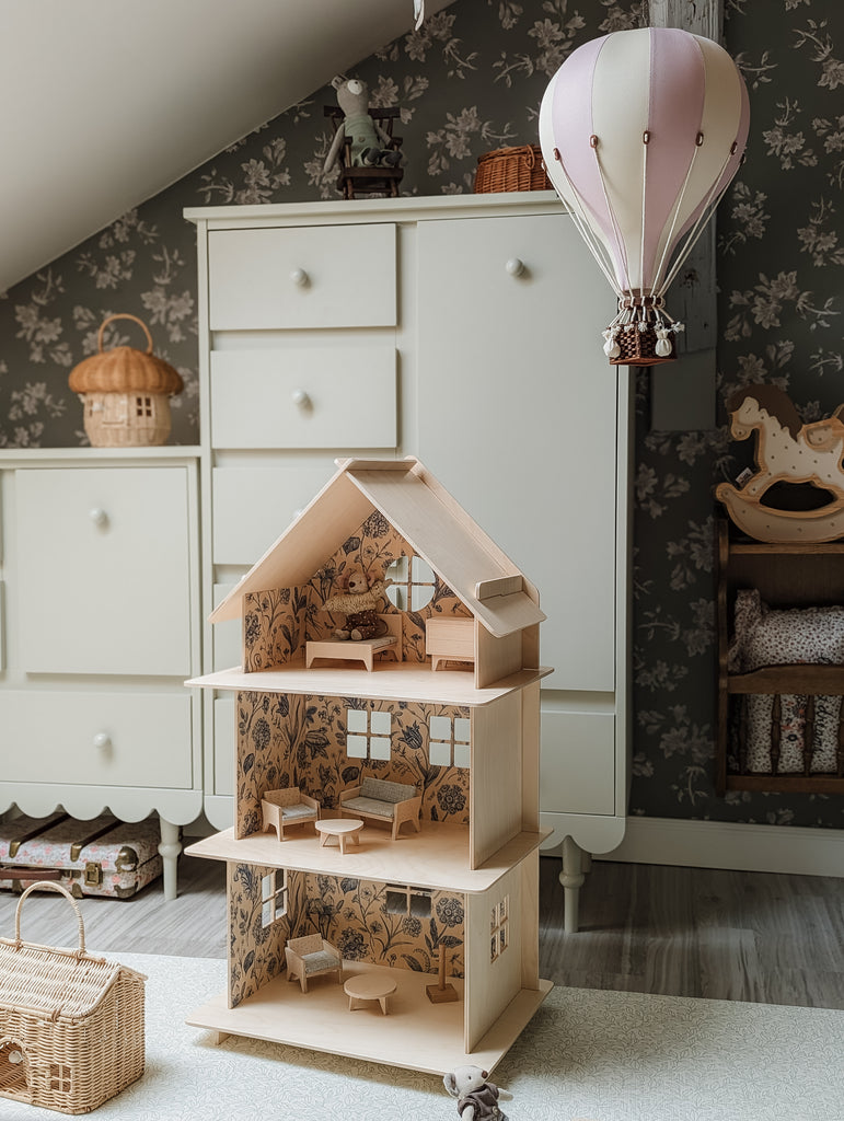 A wooden dollhouse with furniture sits on a rug in a cozy kids’ room decorated with handmade accents. The Hot Air Balloon - Lilac hangs from the ceiling, complemented by white cabinets and a wicker basket in the background.