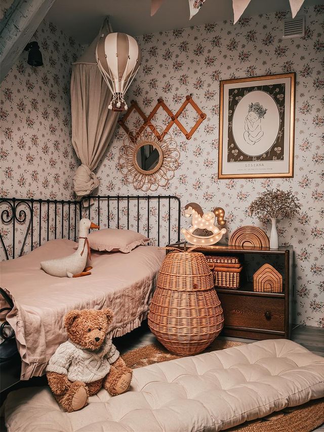 A cozy, vintage-style children’s bedroom with floral wallpaper, pink bedding on a metal bed, wicker baskets, plush toys, and a teddy bear features soft lighting and charming décor like the Hot Air Balloon - Gold.