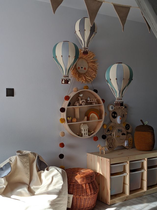 A cozy children's room features a mint green and grey Hot Air Balloon wall decor, a circular shelf with toys, wicker baskets, a soft bean bag, a wooden cabinet with storage boxes, and a pennant banner hanging above.