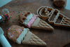 Two ice cream cone-shaped cookies with colorful sprinkles rest on a wooden board, accompanied by the Ice Cream Cutter Set - Eco Playdough Tool and a small container of sprinkles—perfect for inspiring creative sensory play.