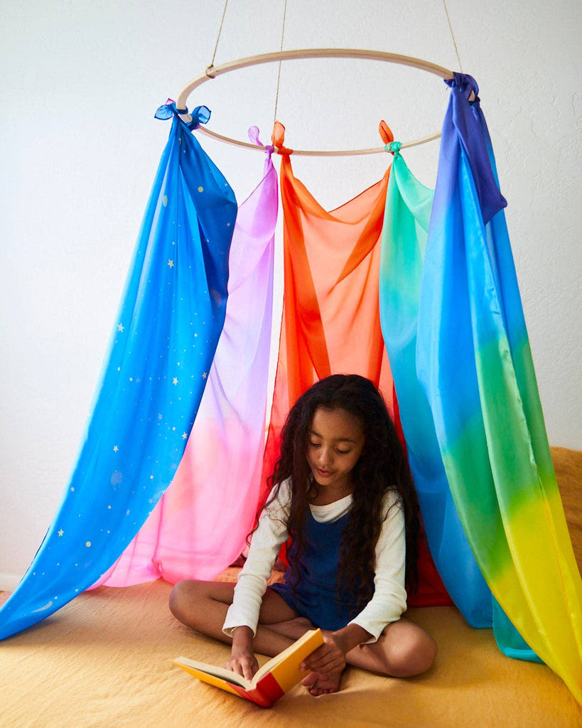 A child sits cross-legged on a bed, engrossed in a book, beneath a vibrant canopy crafted from Sarah's Silk Giant Fire Playsilk. This enchanting setting invites imaginative play as the white-walled room transforms into a world of adventure.