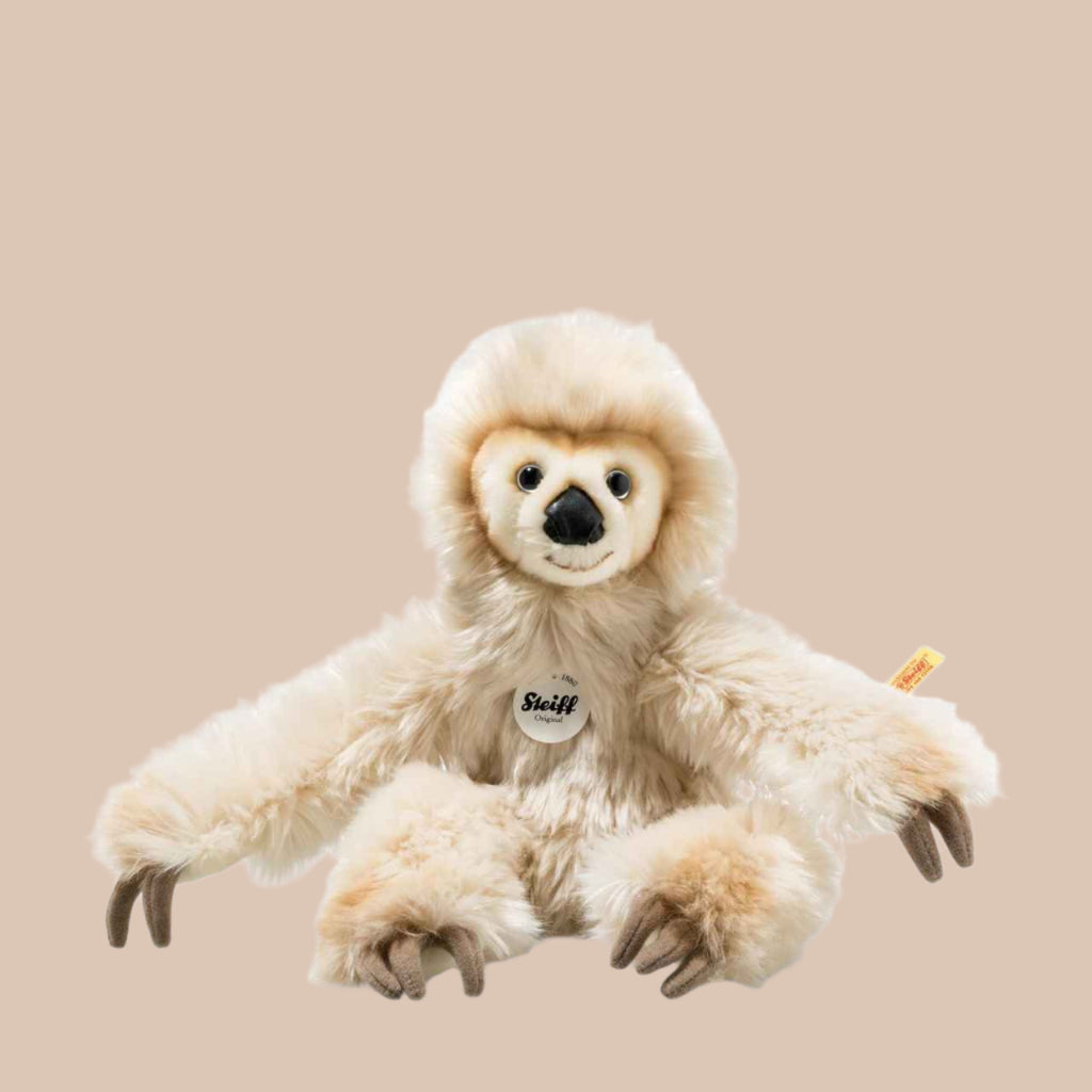 The Steiff Miguel Baby Dangling Sloth Plush Toy, 13 inches, features light brown premium plush fur, long arms, dark claw-like feet, a friendly face, and a small "Steiff" tag on its chest. It is shown sitting against a plain beige background.