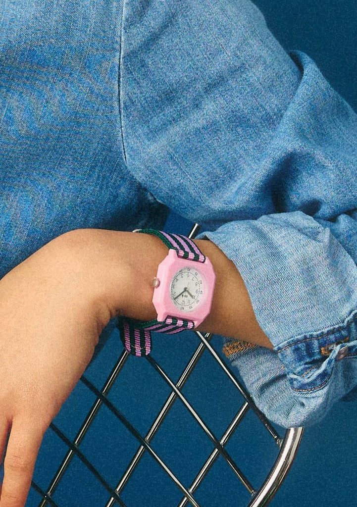 A person in a blue denim shirt rests their arm on a metal chair, showcasing the Mini Kyomo Watch - Lavender Field on their wrist against a blue background.