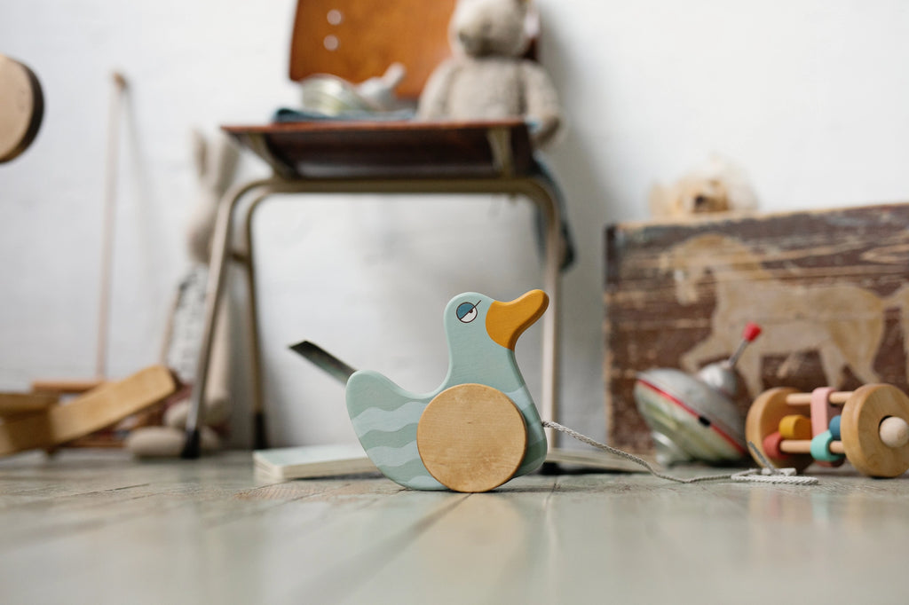 A wooden Duck Pull Toy with wheels on a pastel floor, in a room filled with various vintage toys, creating a nostalgic and playful atmosphere.