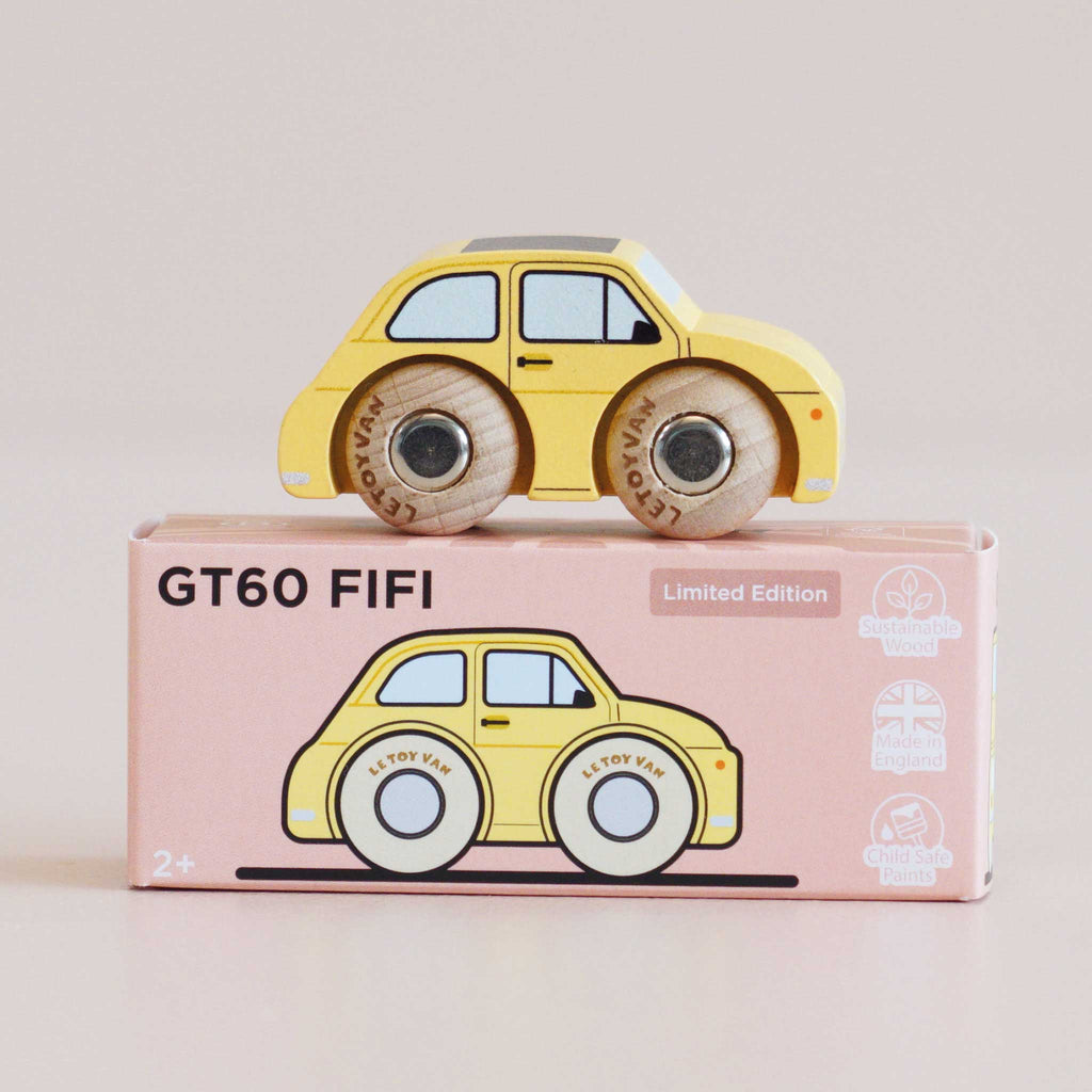 A yellow classic miniature car with wooden wheels rests on a pink box labeled “No. 10 GT60 Fifi | Made in England,” which displays an illustration of the vintage toy car along with various certification icons.