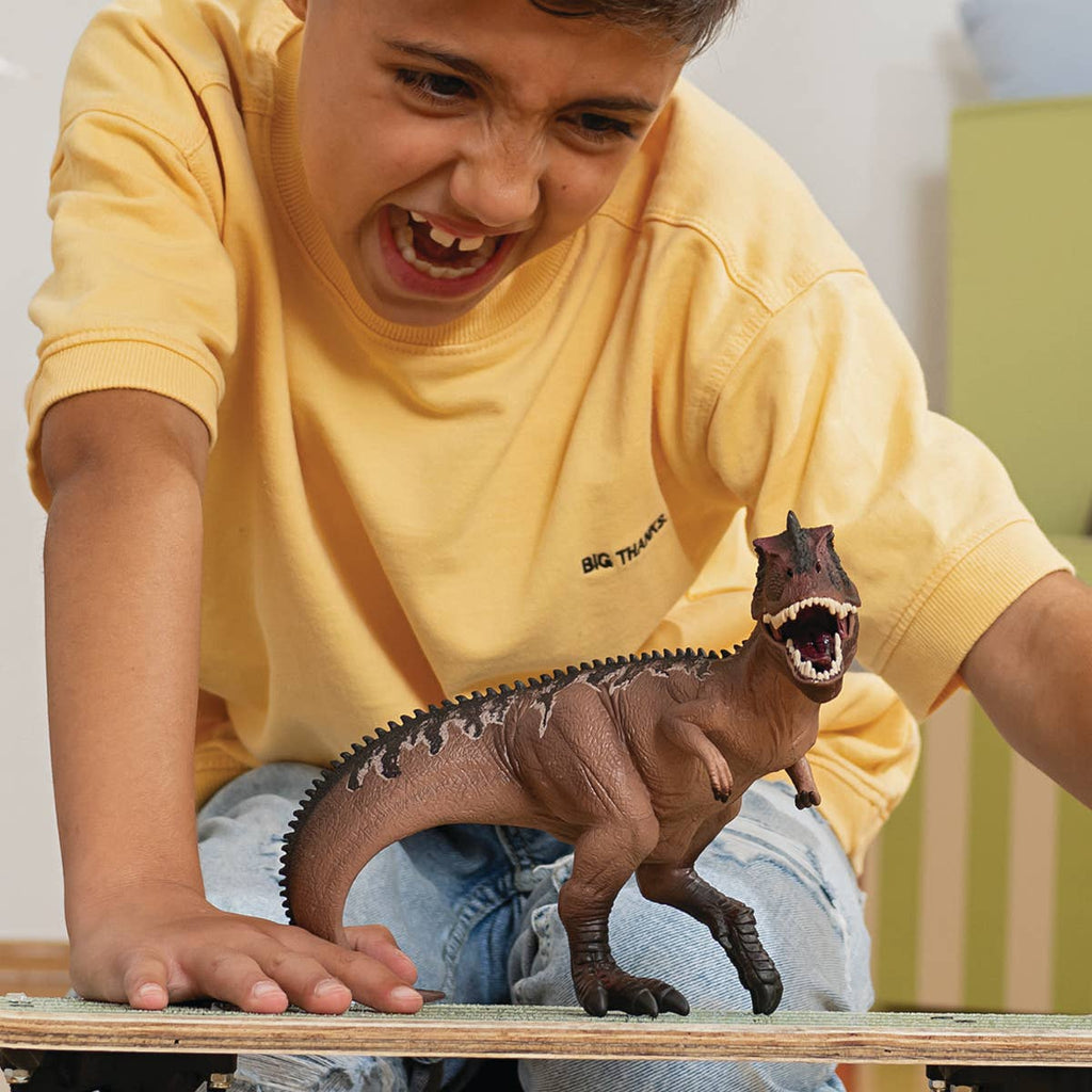 A child in a yellow shirt smiles excitedly while playing indoors with the Schleich Giganotosaurus Large Dinosaur Toy with Movable Jaw on a skateboard.