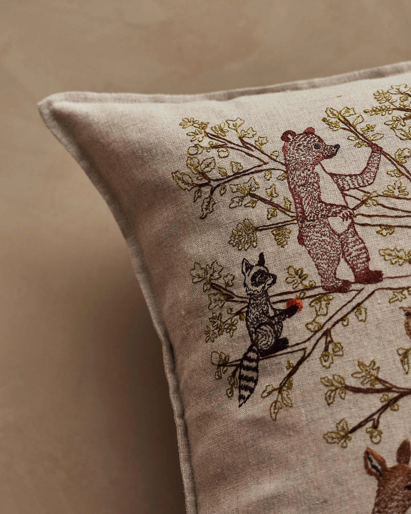 Close-up of the Coral & Tusk Woodland Living Tree Pillow, featuring embroidered forest animals like a raccoon with a red item and a bear among green leafy branches. Ships in one week.