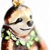 Close-up of a detailed, hand-painted ceramic sloth ornament with a friendly smile. The sloth is adorned with a green leafy garland around its neck and hangs from a 24k gold plated steel chain. The background is plain and white, highlighting the craftsmanship of the Happy Sloth Necklace.